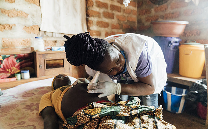 A pregnant woman lies on a bed as a female MSF staff bends over the person with the staff's ear to a cone-shaped stethoscope placed on the woman's stomach.