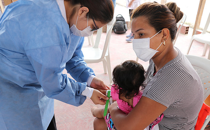 A medical staff puts a green band around the wrist of an infant sitting on the lap of an adult.