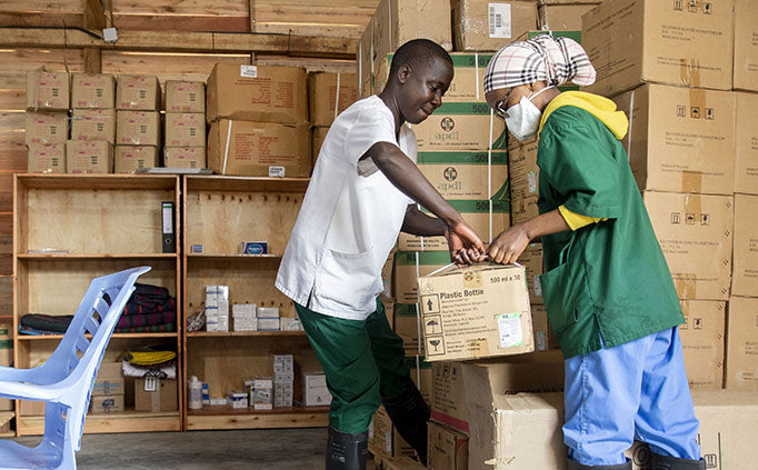 Two MSF staff move cardboard boxes of cholera kits in a room with many other boxes stacked against a wall.