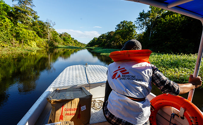 A person wearing a lifejacket sits at the front of a boat as it moves down a river that bisects a forest.