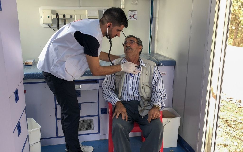 A medical staff uses a stethescope to examine the chest of an elderly person seated in a chair. 