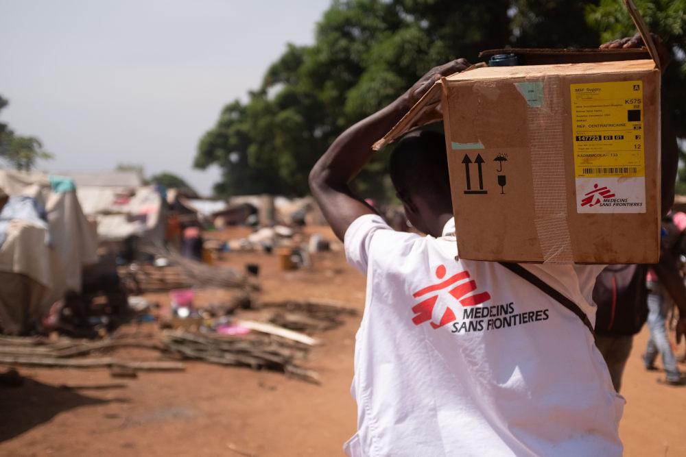 An MSF staff carries on their shoulder a cardboard box of humanitarian relief items. 