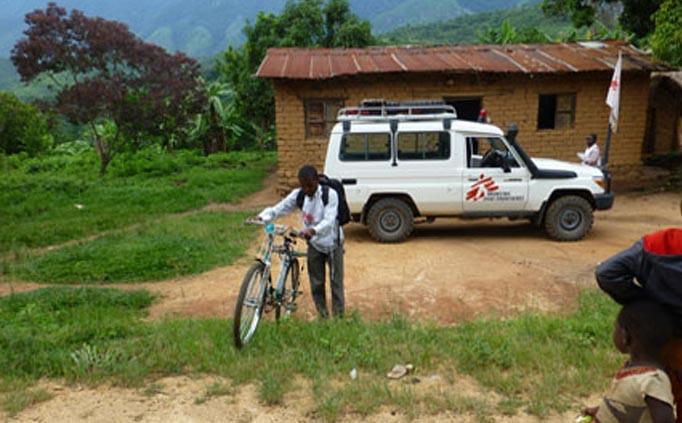 A person walks with a bicycle in front of a rural house and an MSF truck.