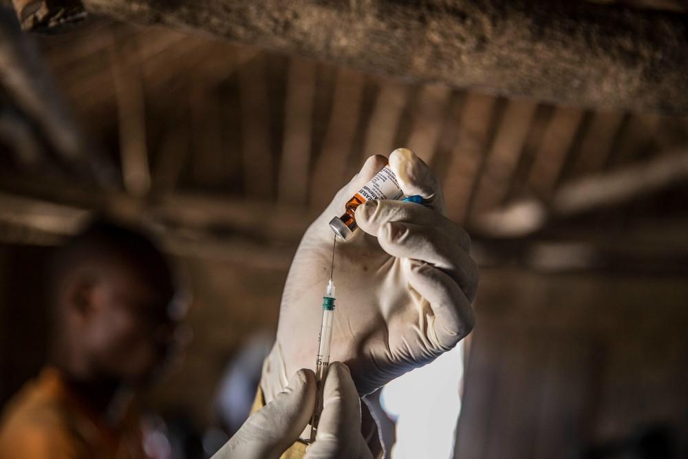 A hand wearing a medical glove puts a syringe into a vial.